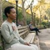 Adult sitting on a bench by a tree-lined path in soft afternoon light with one hand resting gently on their chest, a posture associated with self-compassion practices