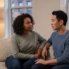 Two people sitting close together on a couch in a warm Manhattan living room, one listening supportively