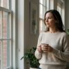 Professional woman pausing by a window with a coffee cup, looking thoughtful in soft morning light in a Manhattan apartment